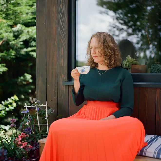 A woman sits outdoors, sipping tea from a patterned cup, wearing a dark green top and a bright orange skirt, beside a wooden-clad building with a lush garden and reflective window in the background.